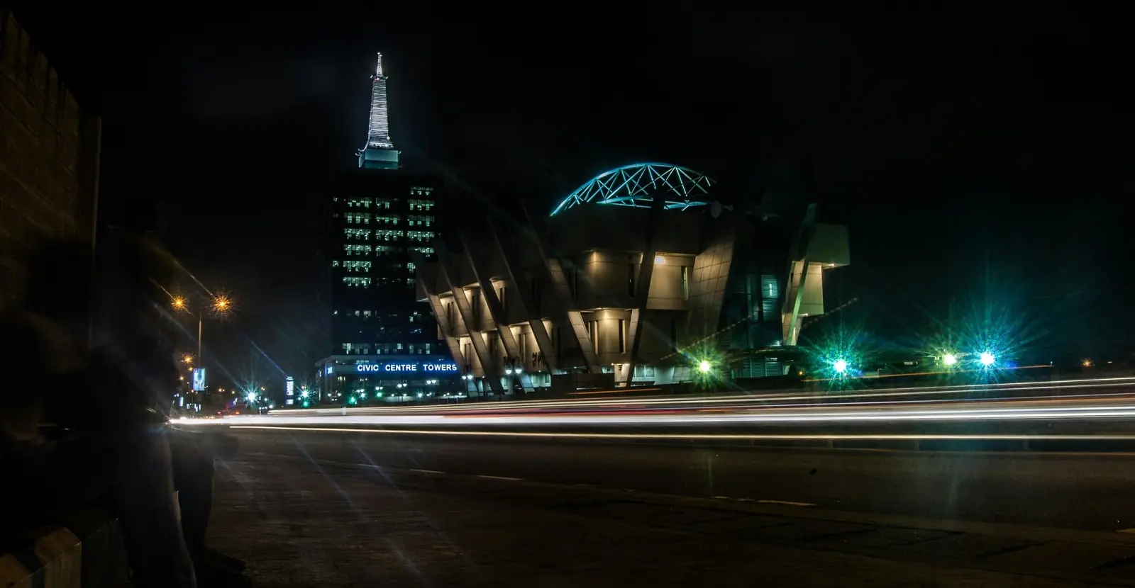 Lagos at night: a lit office tower and civic building beside a road, with light trails from passing traffic.